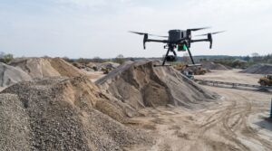 Realistyczne zdjęcie z poziomu ziemi przedstawiające profesjonalny, duży dron przemysłowy (quadcopter) w trakcie lotu nad rozległym terenem żwirowni lub placu budowy. Dron jest czarny, wyposażony w widoczne czujniki i kamerę zamontowaną pod spodem, a jego cztery śmigła są w ruchu. Unosi się w powietrzu na tle ogromnych, nieregularnych hałd kruszywa o różnej wielkości i barwach – od drobnego, jasnego piasku po ciemniejszy, gruboziarnisty żwir i kamienie. Teren wokół jest piaszczysty i pokryty śladami ciężkich pojazdów. W tle, na rozległym, otwartym obszarze pod jasnym, lekko zachmurzonym niebem, widoczne są maszyny budowlane, takie jak koparki i ładowarki, a także taśmociągi i naturalna roślinność (drzewa) na horyzoncie. Kadrowanie ukazuje techniczną pracę drona mierzącego objętość zgromadzonego materiału w naturalnym, dziennym oświetleniu.